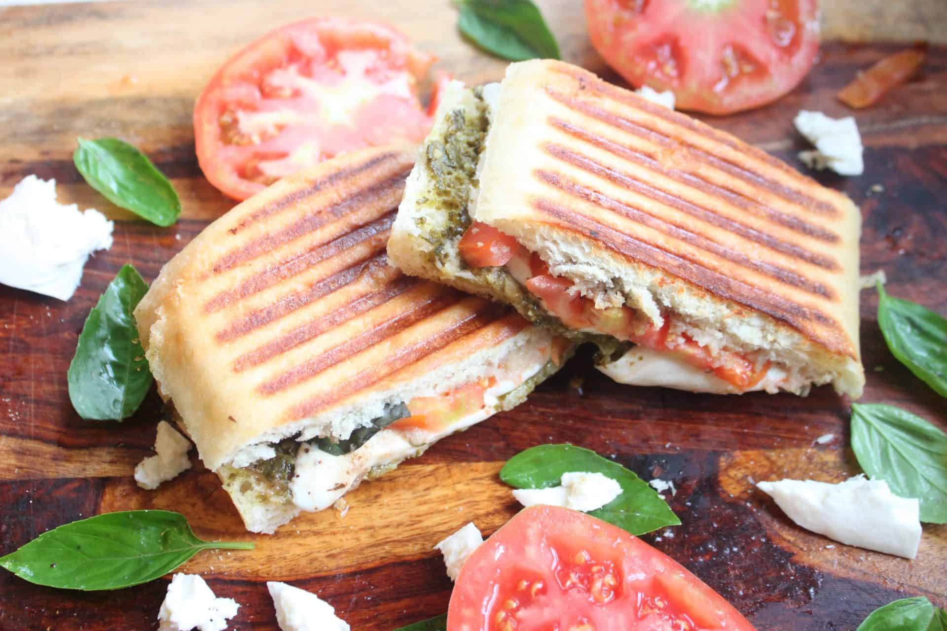 Sliced Grilled Caprese Sandwich Sitting on a Cutting Board Surrounded by Tomato Slices, Basil Leaves, and Mozzarela
