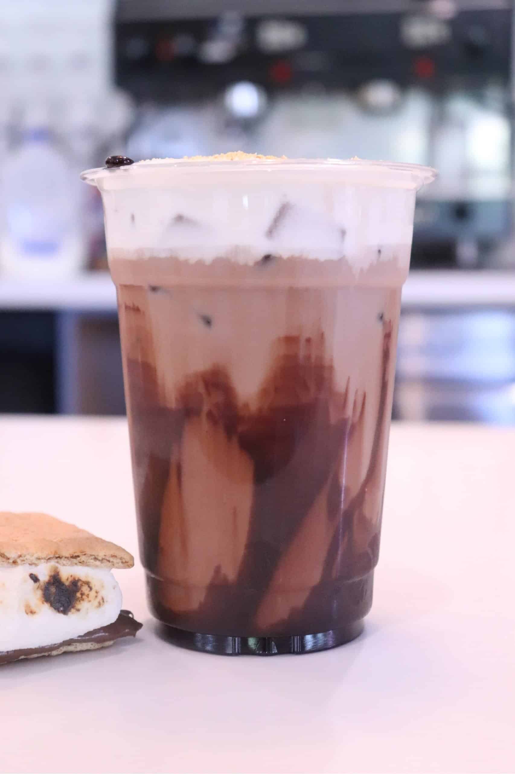 Iced smores latte topped with whipped foam, next to a toasted marshmallow and chocolate graham cracker sandwich in the foreground.