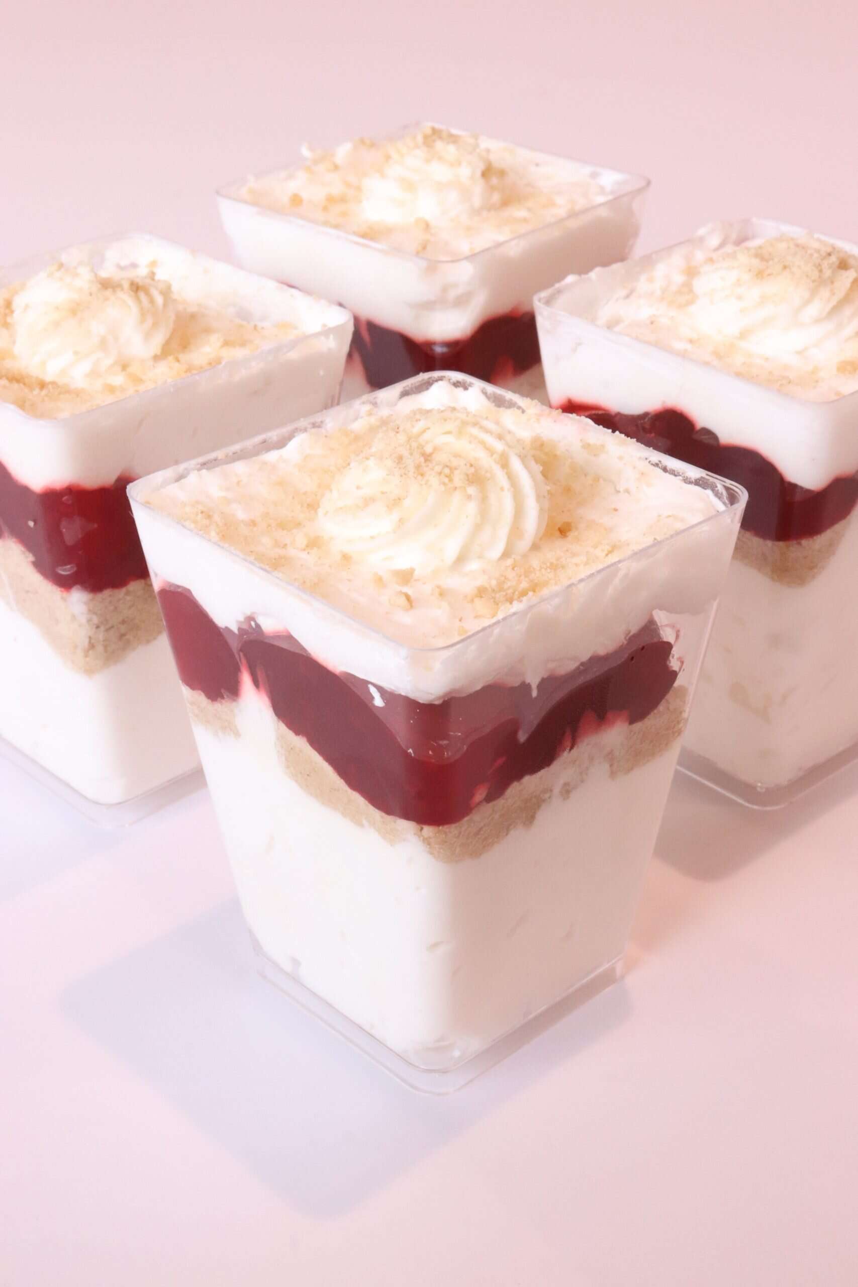 Four strawberry cheesecake trifles layered with creamy filling, strawberry sauce, and graham crumbs, all served in clear square cups.