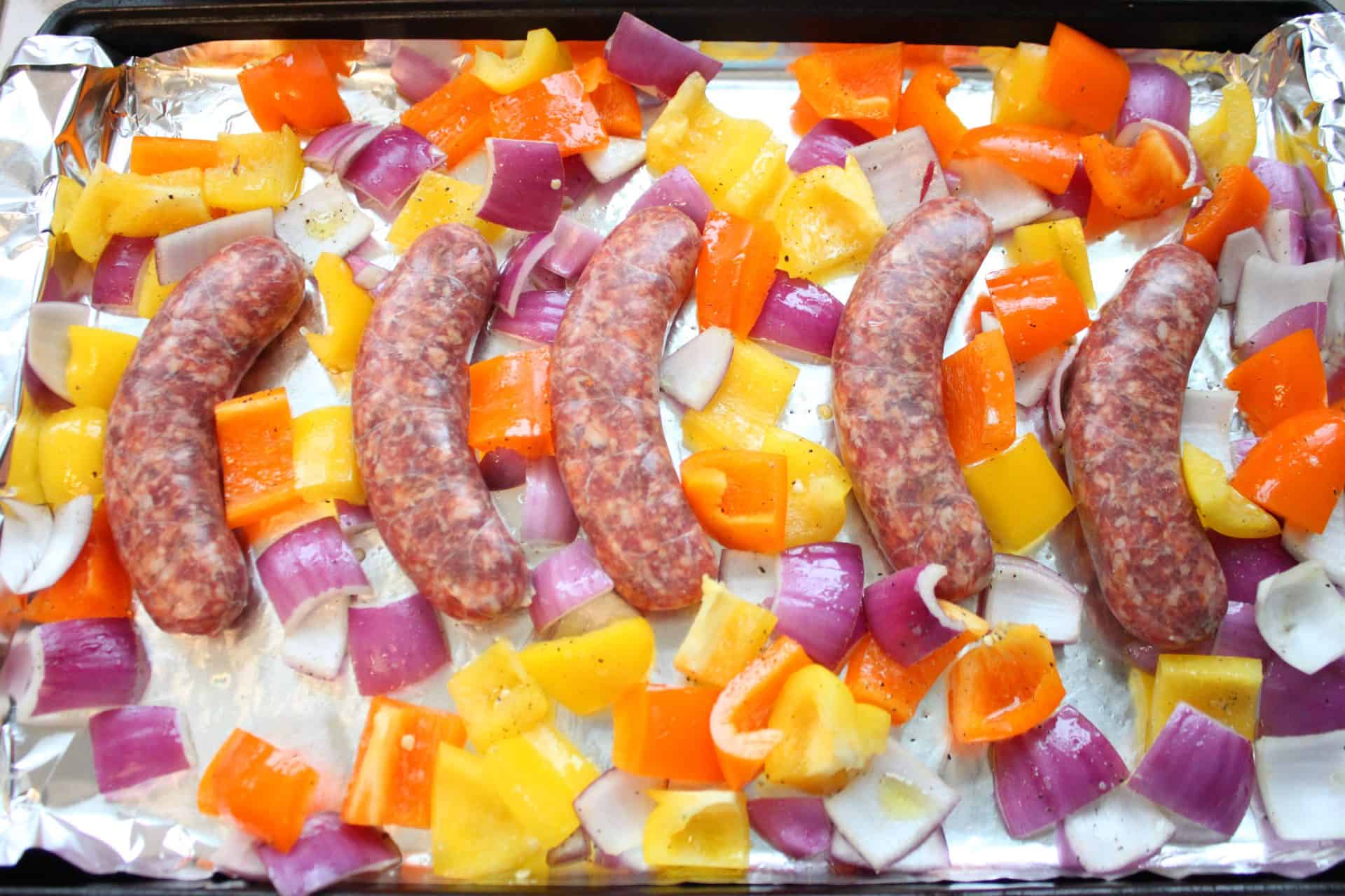 Uncooked Italian sausages arranged on a sheet pan with chopped bell peppers and red onions, ready to roast for hoagie sandwiches.