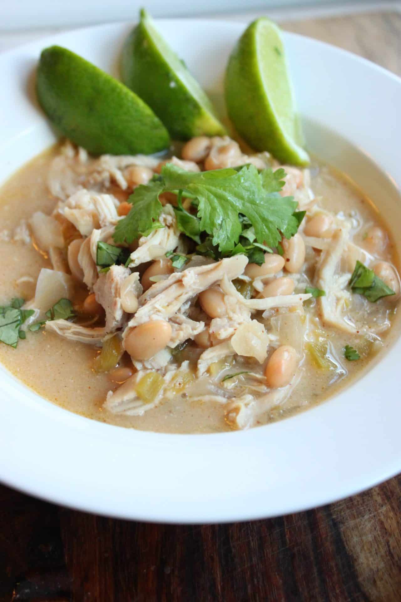 A vertical shot of Healthy Crockpot White Chicken Chili in a white bowl, garnished with cilantro and lime wedges.