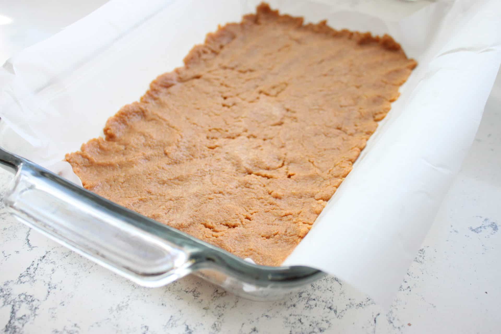 Pressed graham cracker crust for Coconut Pecan Bars in a glass baking dish lined with parchment paper, ready for layering.
