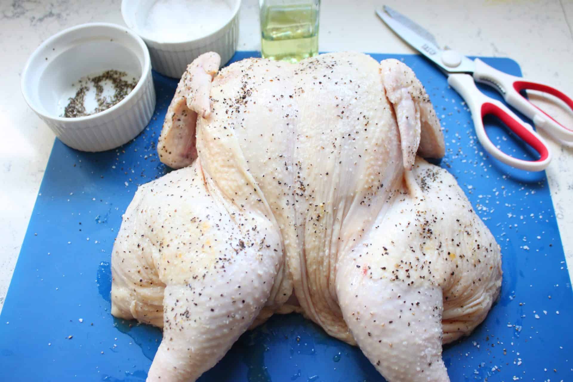 Raw spatchcock chicken on a blue board, rubbed with olive oil, kosher salt, and pepper as part of the seasoning step before baking.