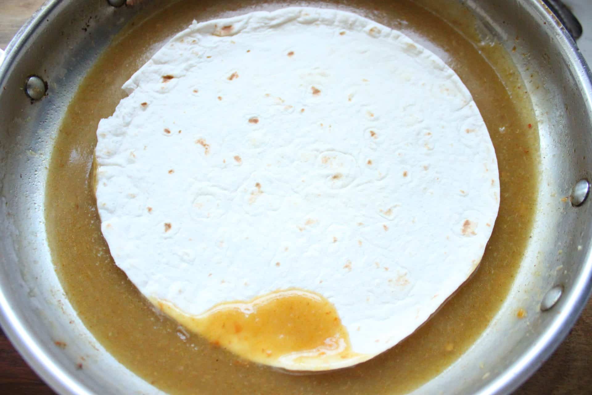 How to Make Flavor-Packed Green Chile Chicken Enchiladas at Home 7 A flour tortilla dipped in a pan of green chile sauce, preparing it for rolling into green chile chicken enchiladas.
